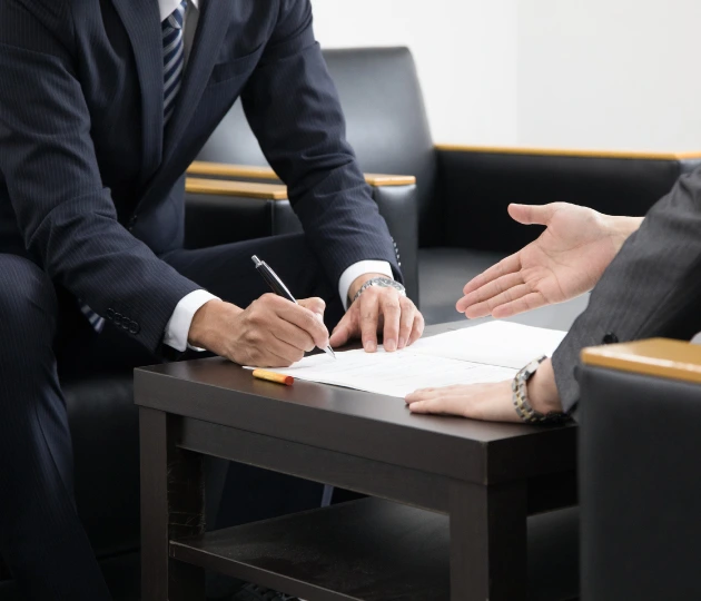 Close-up of a professional in a dark suit signing a document on a table, with another person gesturing towards the paperwork.